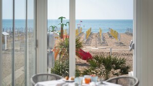 A beach view from a room with a table set for breakfast, featuring a view of the beach with umbrellas and chairs.