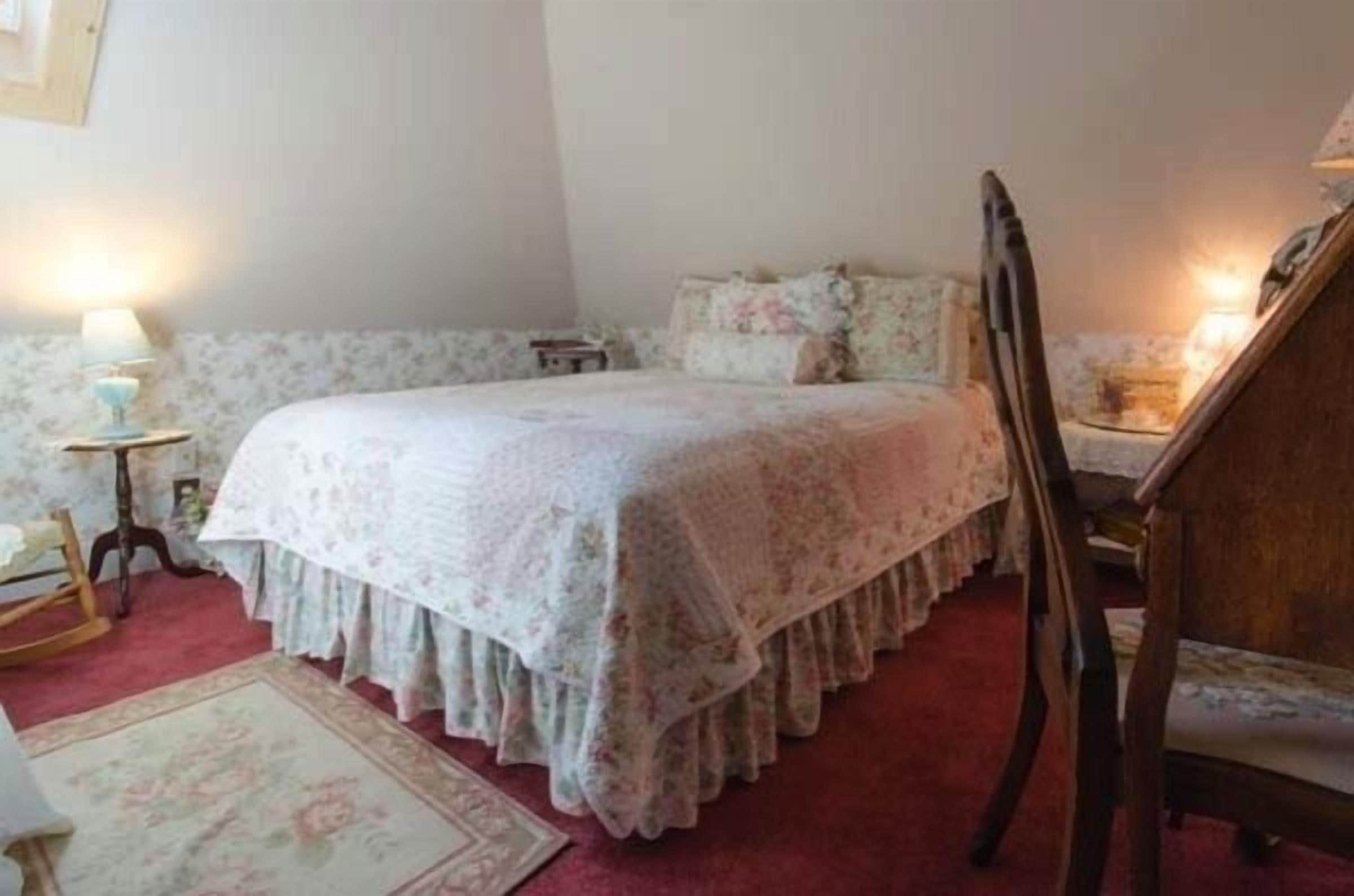 Una camera da letto con una coperta dal motivo floreale, una poltrona a dondolo in legno, un comodino con una lampada e un tappeto con fiori.