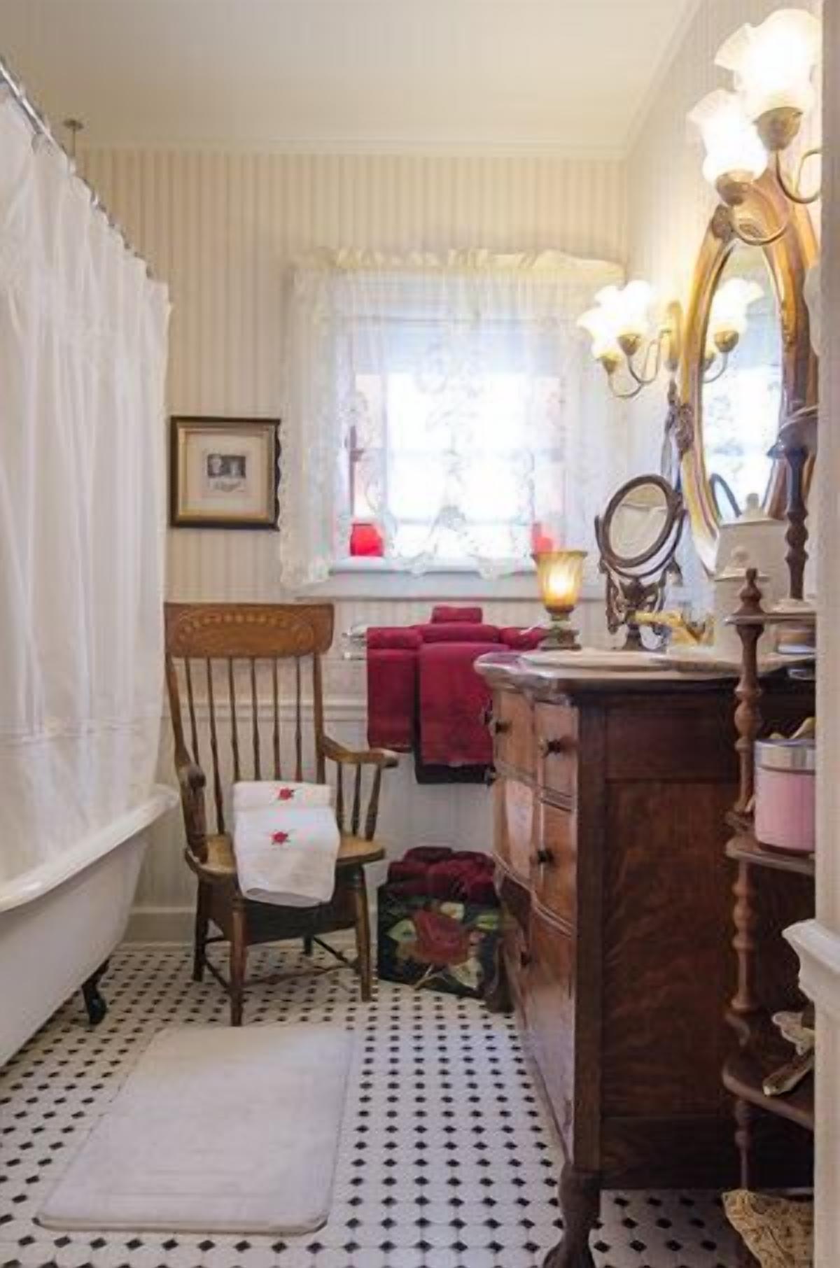 A bathroom with a bathtub, a wooden chair, a vanity with a mirror, and a window with curtains.
