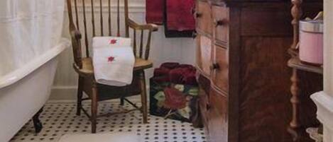 A bathroom with a bathtub, a wooden chair, a vanity with a mirror, and a window with curtains.