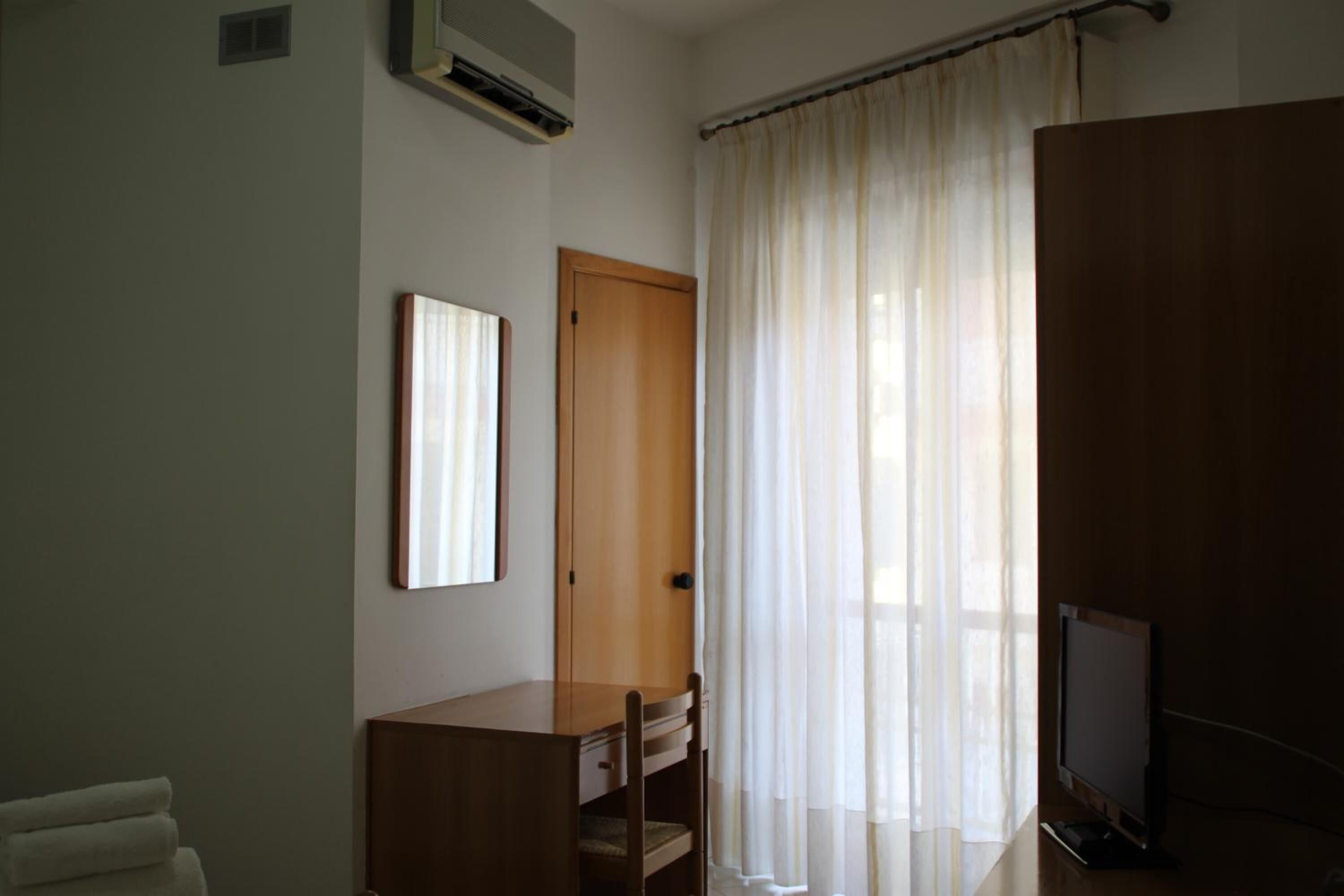 A room with a wooden desk, a television, and a window with sheer curtains.