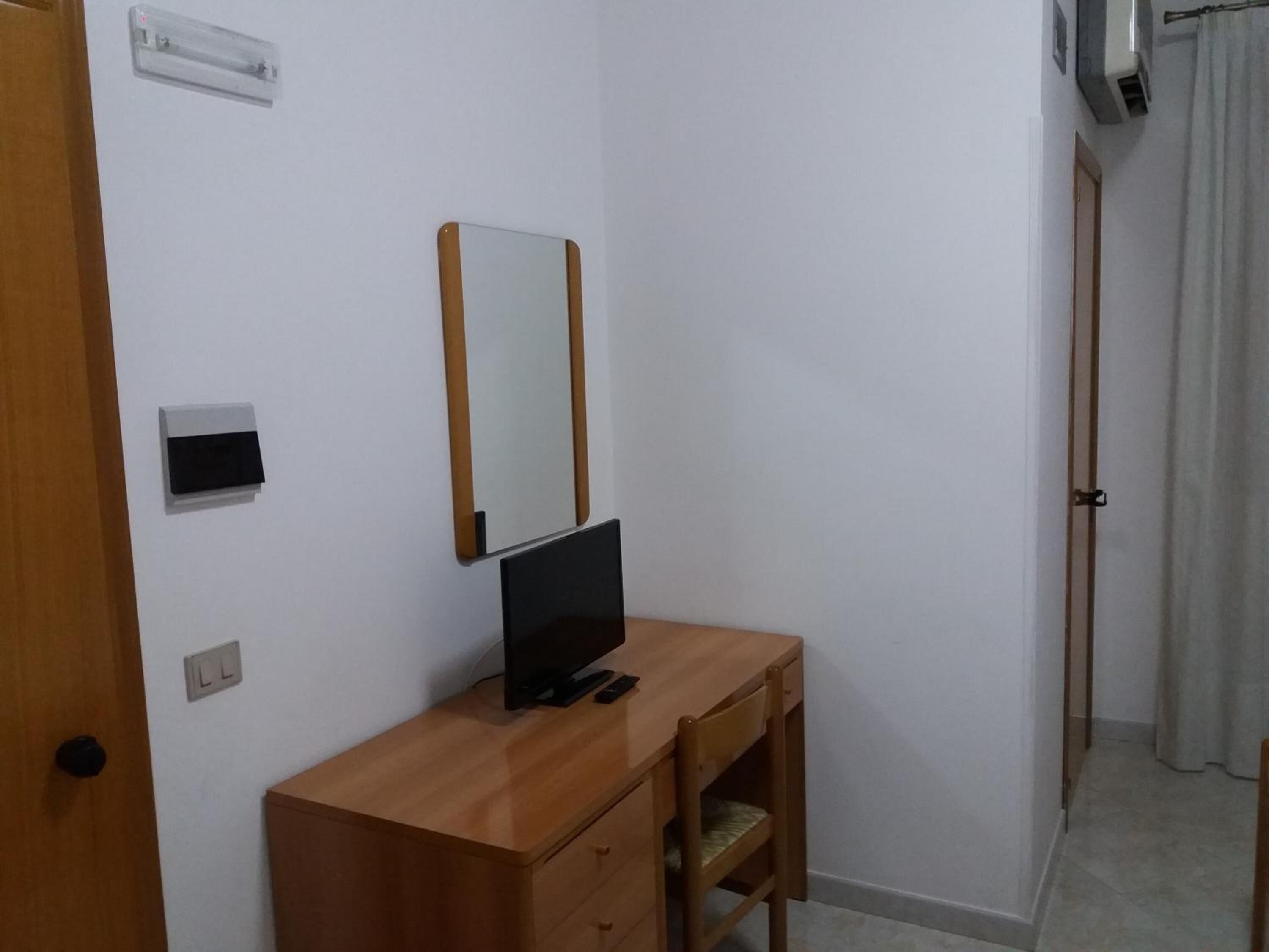 A small, simple room with a wooden desk, a flat-screen TV, a mirror, and a door.