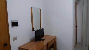 A small, simple room with a wooden desk, a flat-screen TV, a mirror, and a door.
