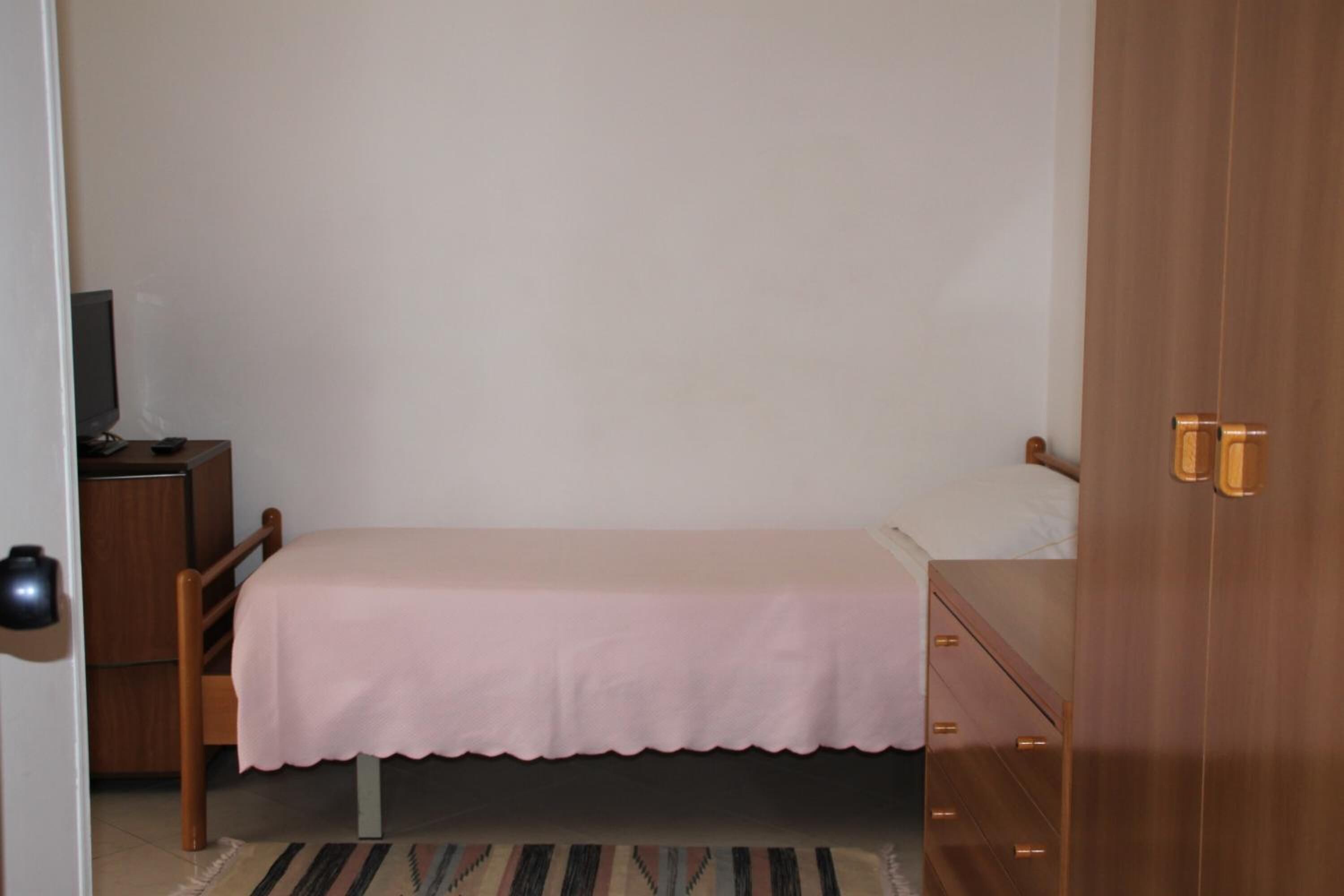 Una habitación pequeña con una cama individual, un armario de madera, una mesita de noche y un televisor sobre un soporte.