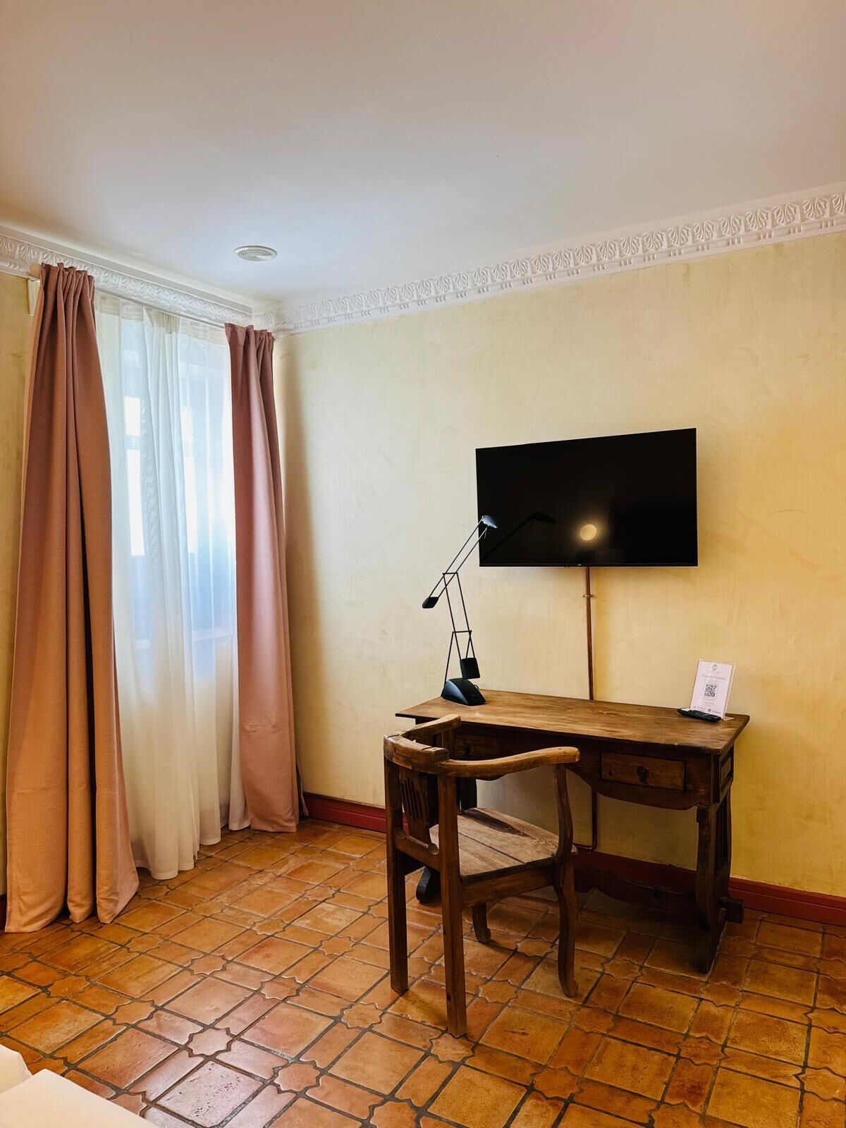 Een kamer met een houten bureau, een stoel, een aan de muur bevestigde televisie en een raam met gordijnen.