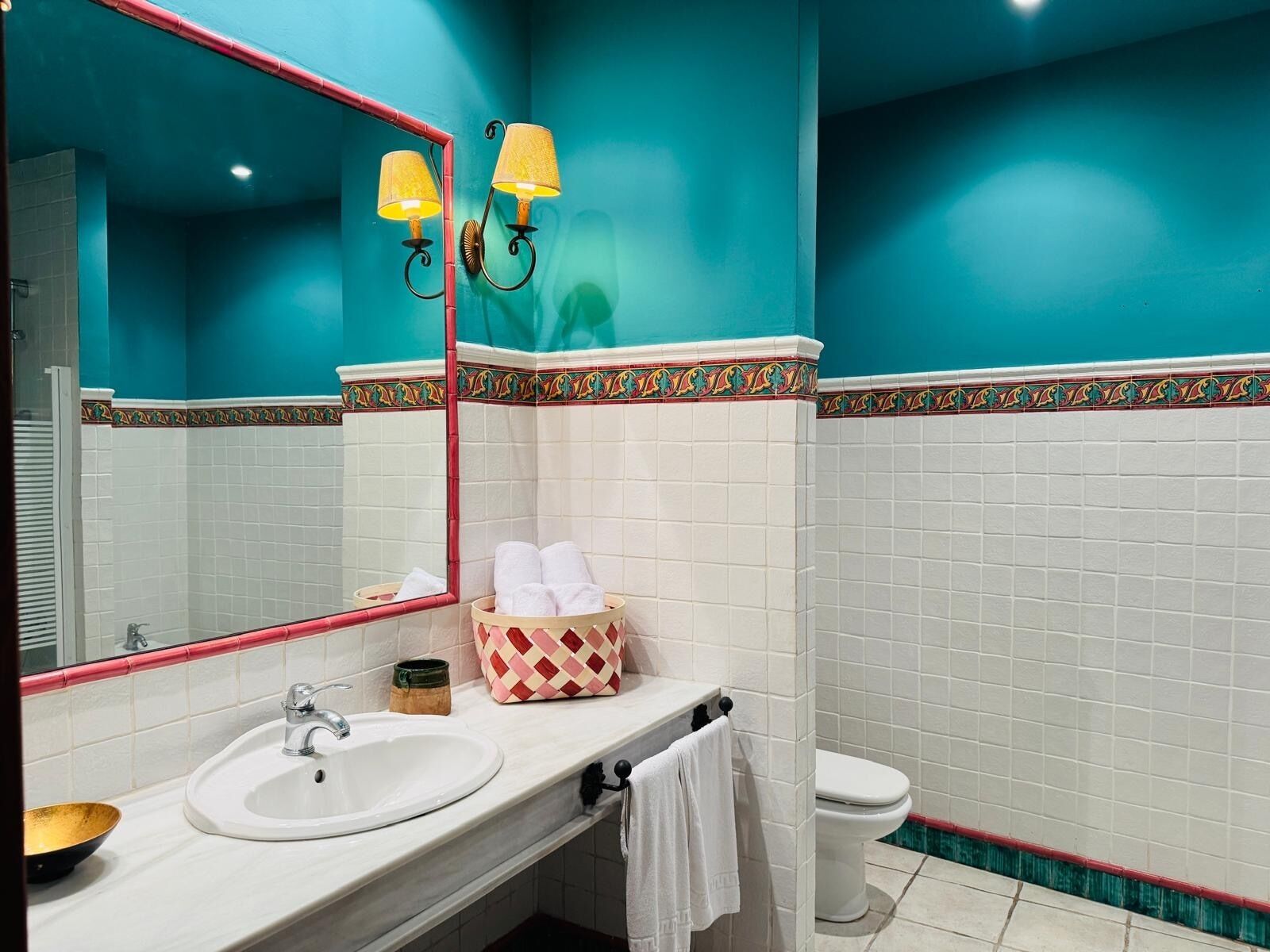 Un baño con lavamanos blanco, espejo con marco rojo, inodoro y un detalle decorativo en la pared.