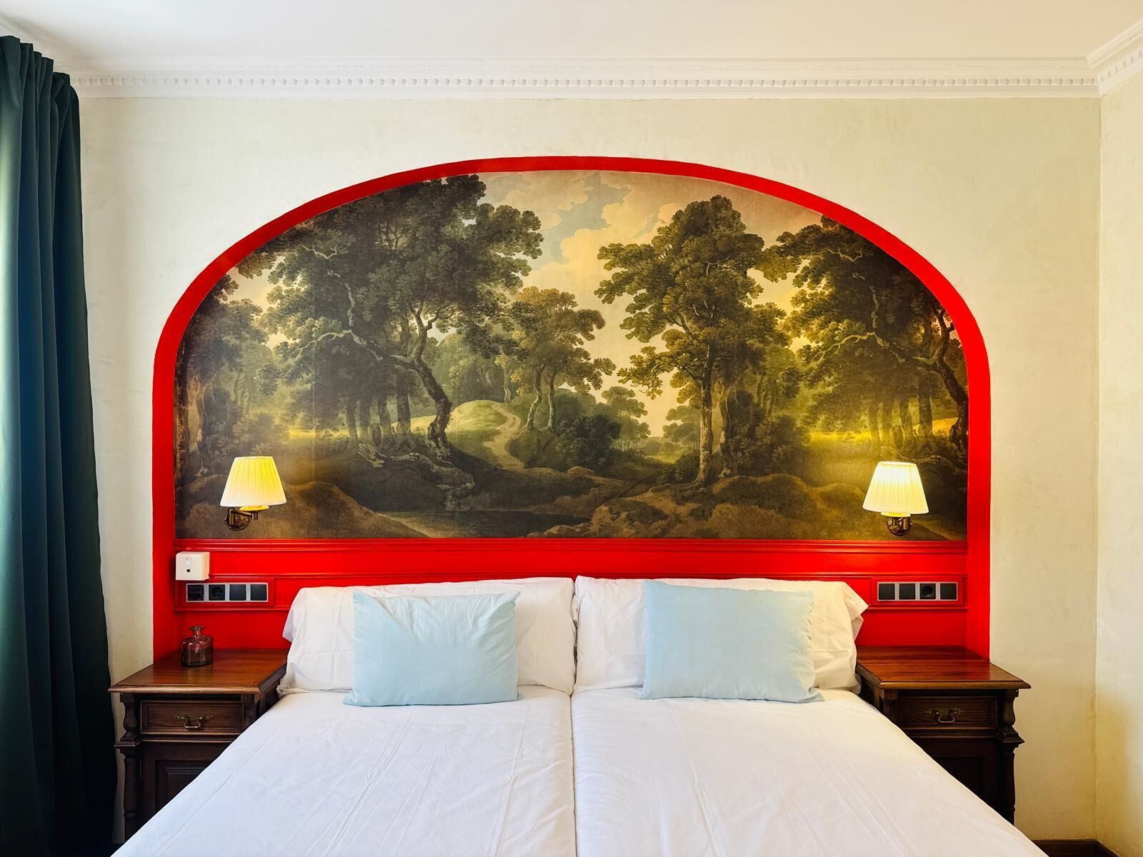 Quarto de hotel com uma cama grande, duas luminárias de cabeceira e um quadro com moldura vermelha retratando uma paisagem florestal na parede.