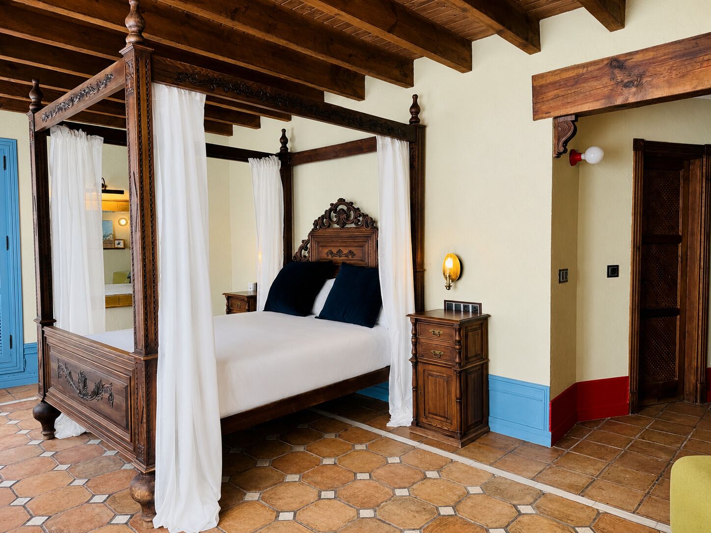 Una cama con dosel y cuatro columnas, una mesita de noche con lámpara, y un suelo de baldosas.