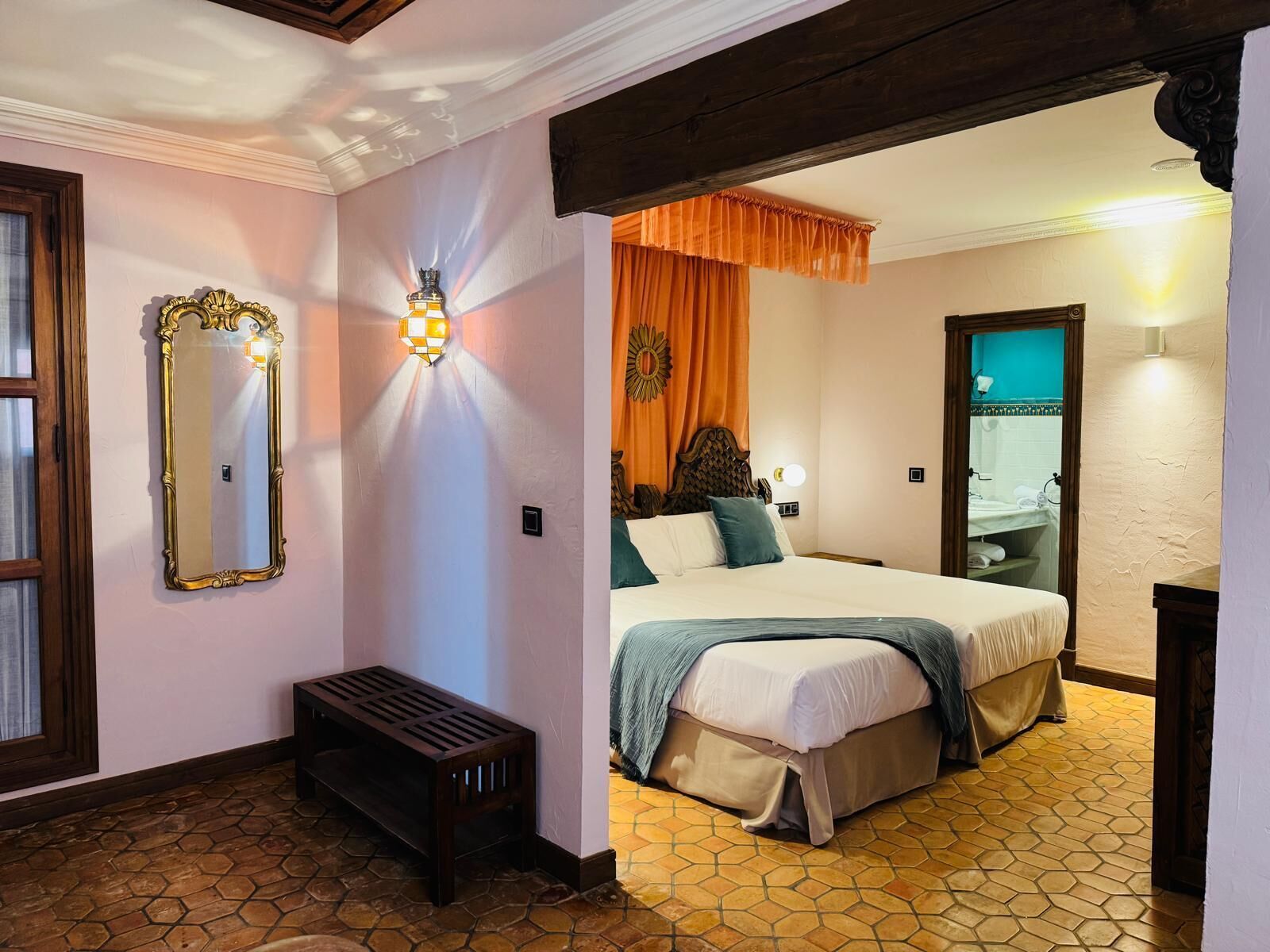 A hotel room with a bed, a bench, a mirror, and a bathroom visible through an open door.