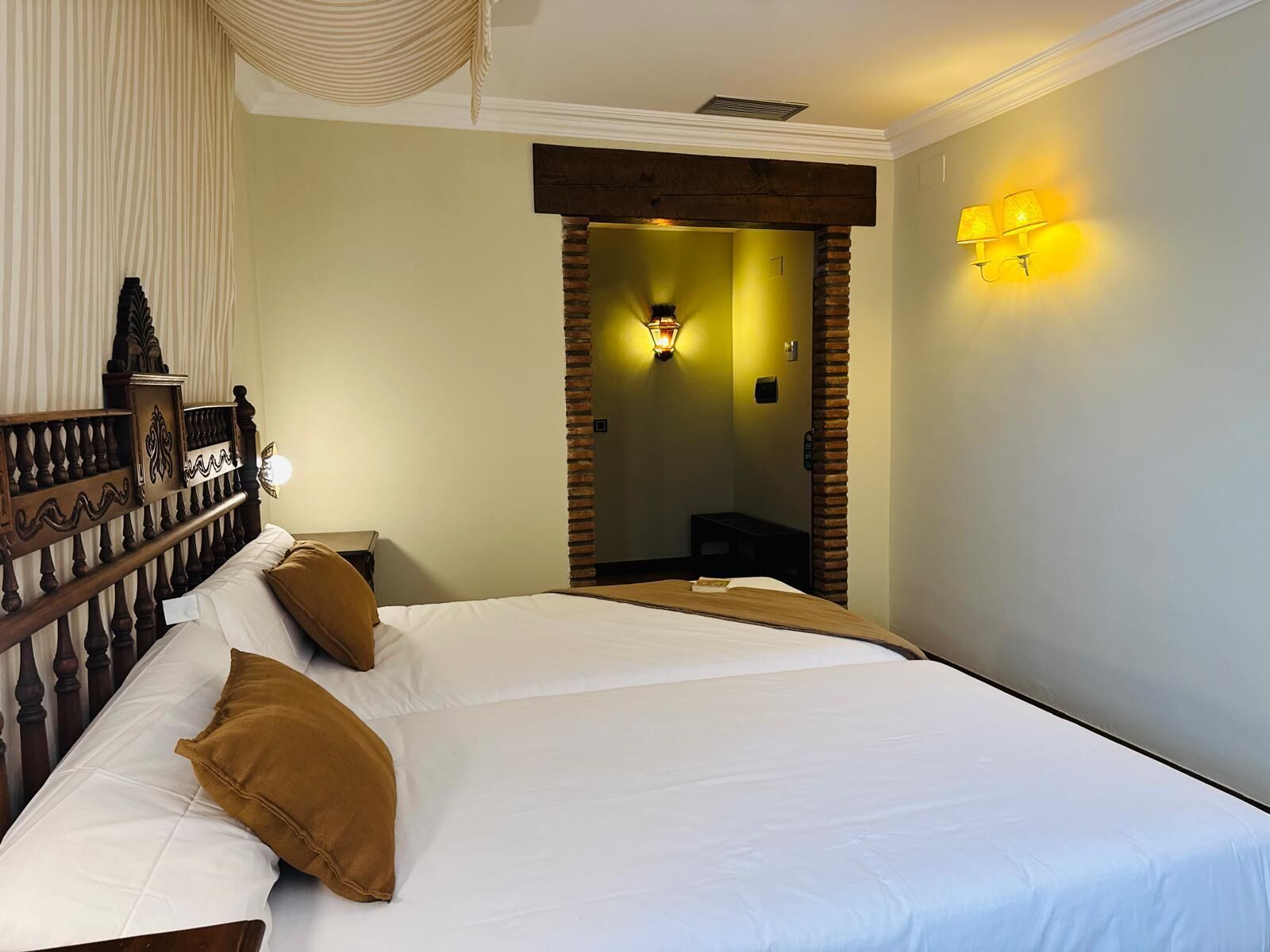 A hotel room with a large bed, a wooden headboard, a nightstand, and a wall-mounted lamp.