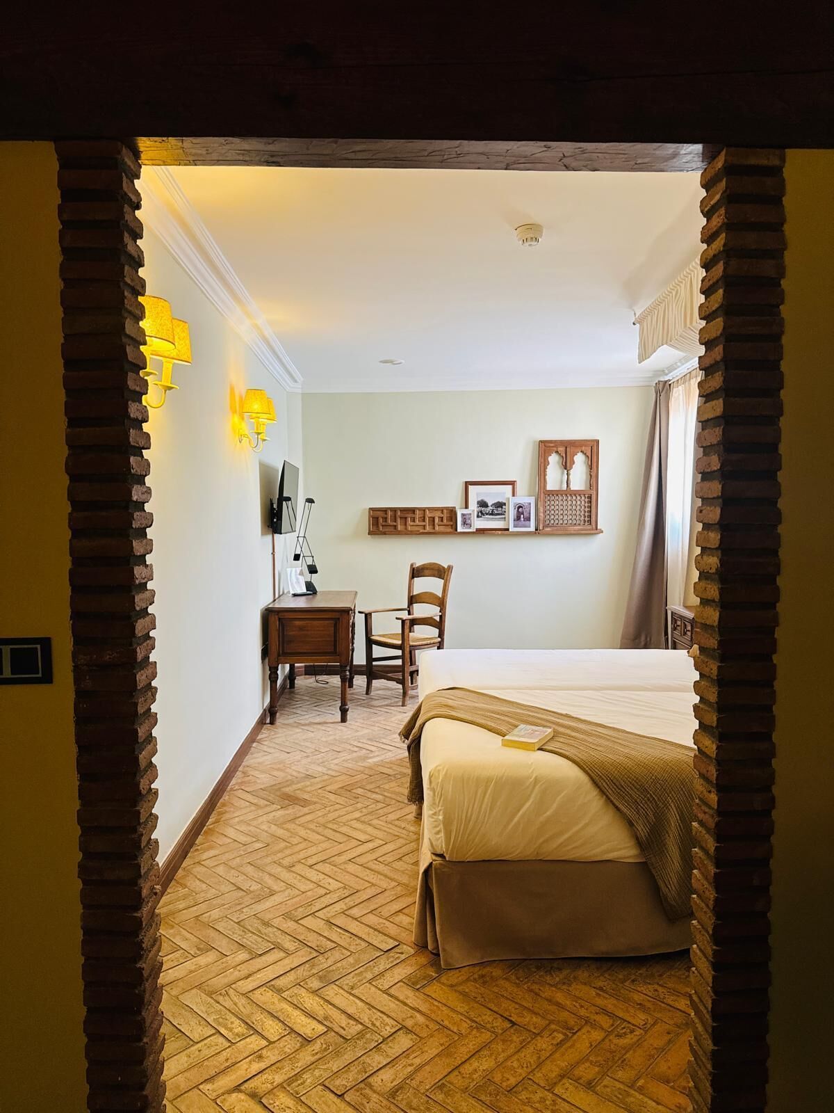 Une chambre d’hôtel avec un parquet en bois disposé en chevrons, un lit recouvert d’une courtepointe beige, un bureau avec une chaise et des lampes fixées au mur.