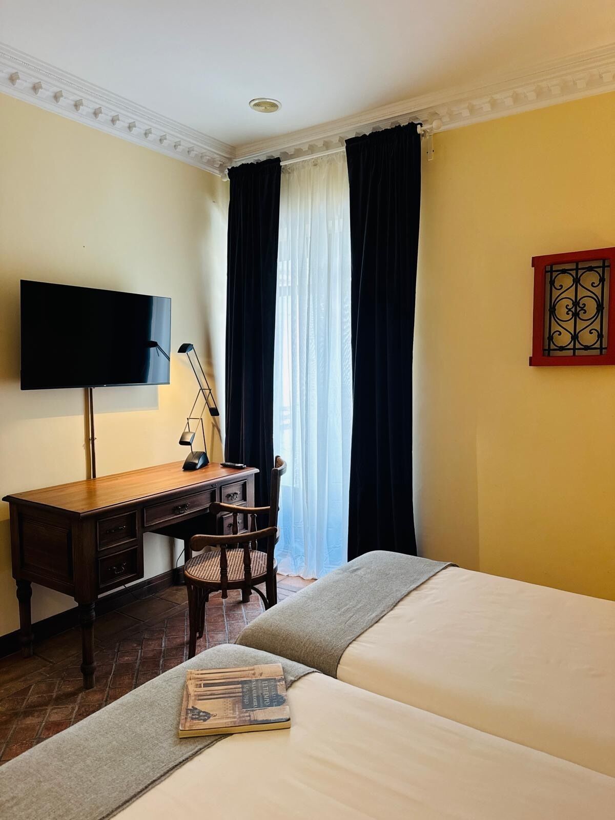 A hotel room with a bed, a desk with a chair, a television, and a window with curtains.