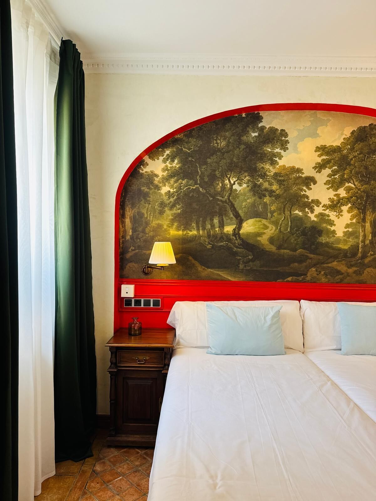 Una habitación de hotel con una cama, una mesita de noche, una lámpara y un cuadro grande de paisaje en la pared.