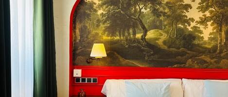 Una habitación de hotel con una cama, una mesita de noche, una lámpara y un cuadro grande de paisaje en la pared.