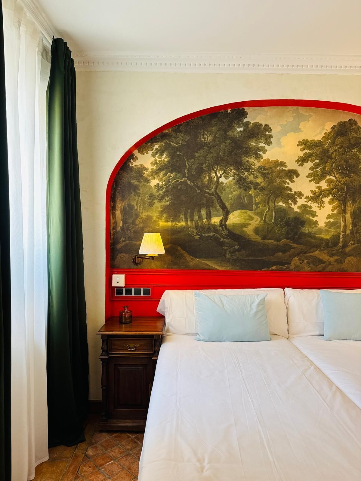 Habitación de hotel con cama, mesita de noche, lámpara y un cuadro grande con un paisaje en la pared.