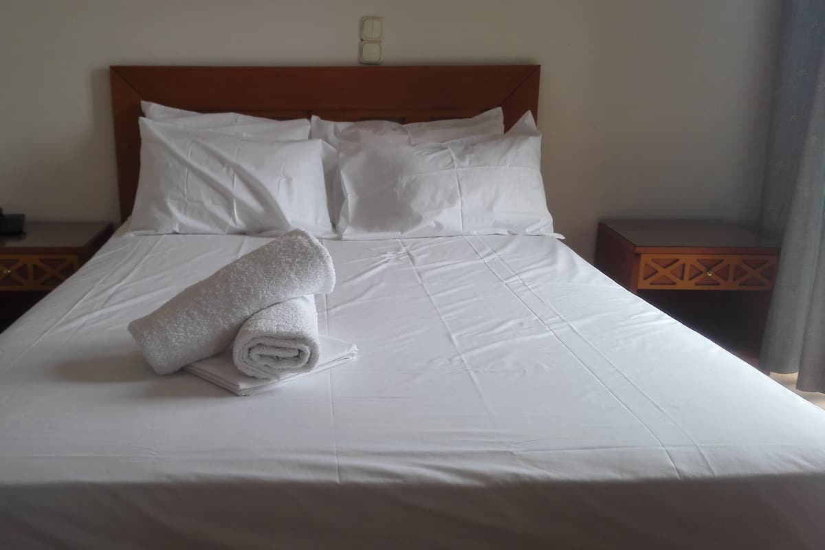 A neatly made bed with white linens and a rolled towel on the bed. There are two nightstands with a phone and a small item on each.