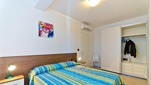 Ein Schlafzimmer mit Bett, Nachttisch, Lampe, Klimaanlage und einem Kleiderschrank mit Kleidung.