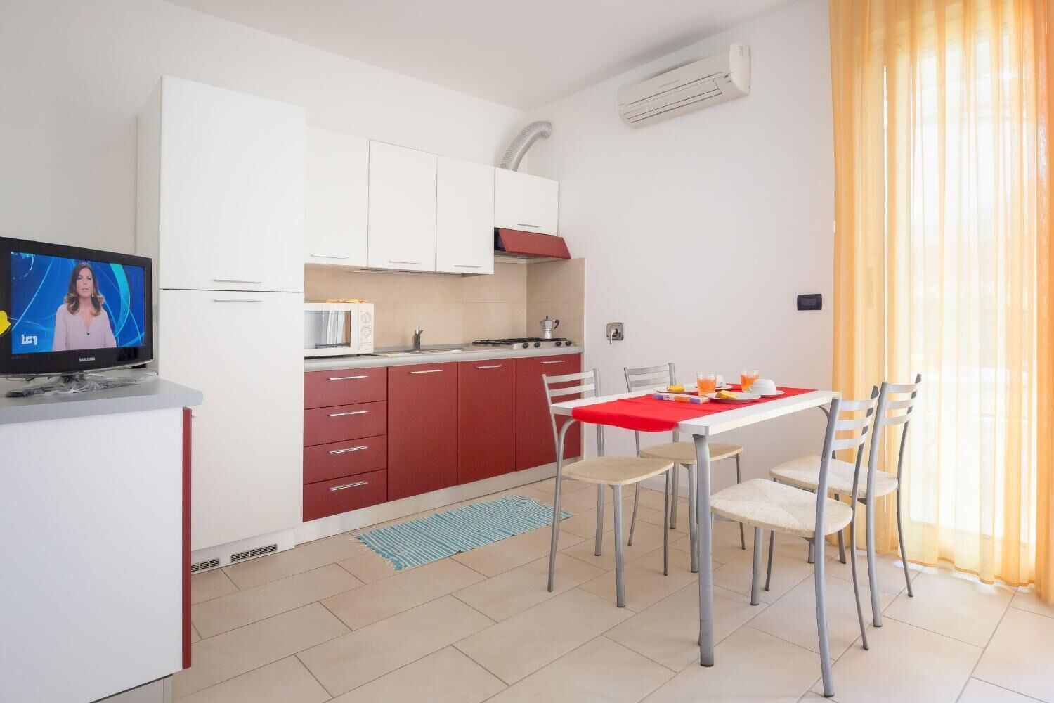 Une cuisine moderne avec des meubles blancs et rouges, un four à micro-ondes et un coin repas agrémenté d’une nappe et de chaises rouges.