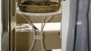 Un balcón con una cesta de mimbre que contiene flores, una mesa blanca y vistas a un pueblo blanco con una construcción similar a un castillo en una colina.