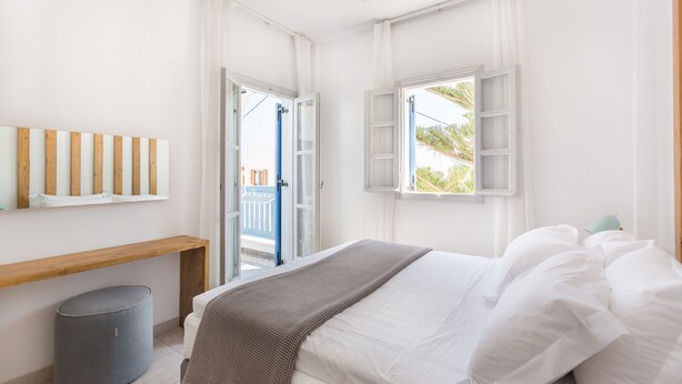A bedroom with a bed, a wooden bench, a grey ottoman, and a window with curtains. - Fanouris Condo by Meraki Collection Adults Only (18+) accommodation (Santorini)