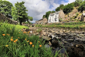 A stone bridge over a stream, surrounded by lush greenery and historic buildings. - Plas Tan y Graig (Caernarfon)