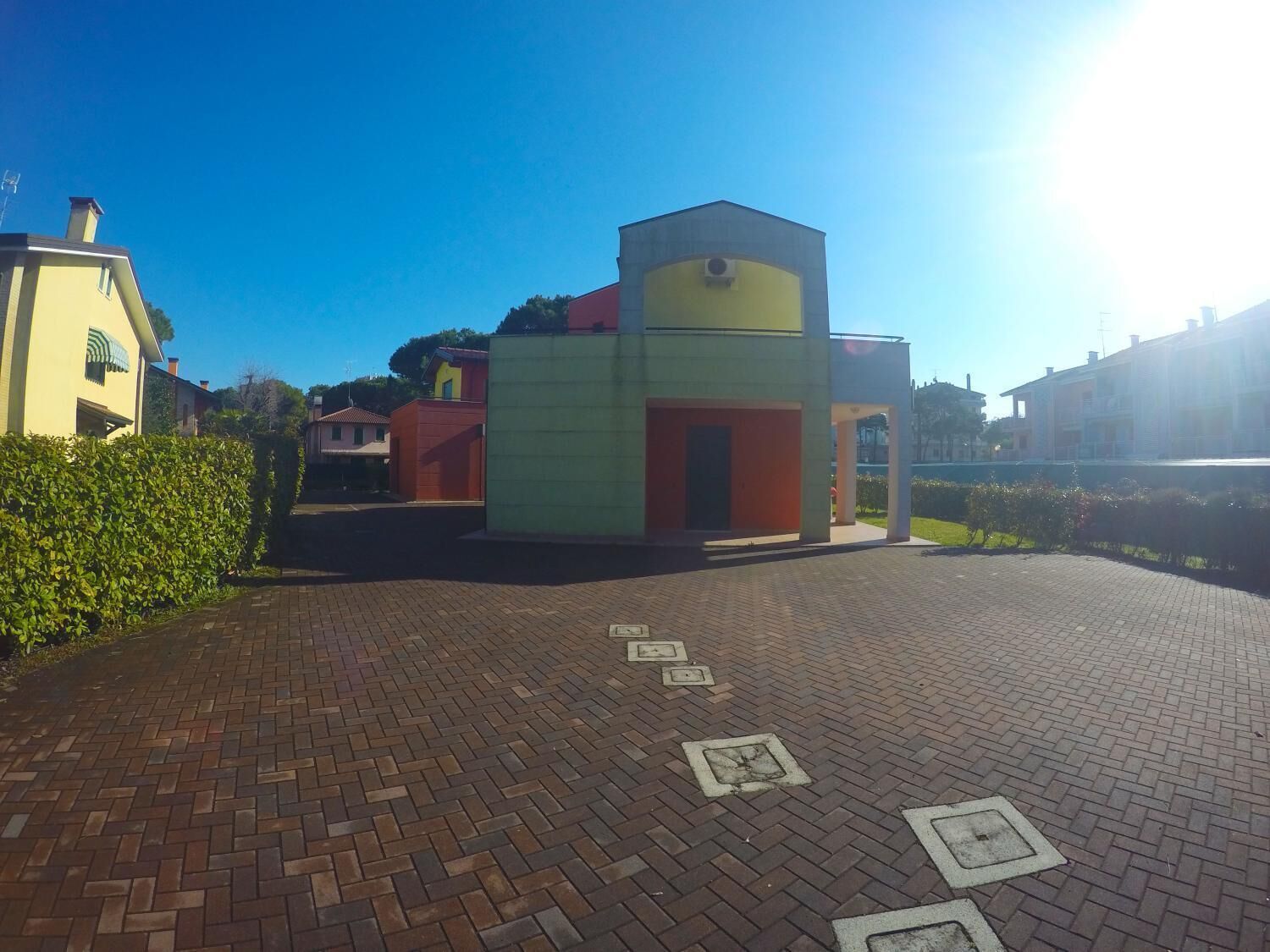 A paved courtyard with a small building, a hedge, and a clear blue sky.