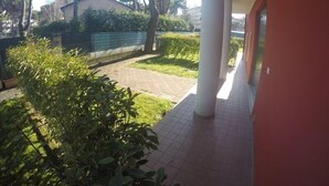 A covered outdoor area with a tiled walkway, greenery, and a building with a red facade. - Residence Le Piramidi (Caorle)