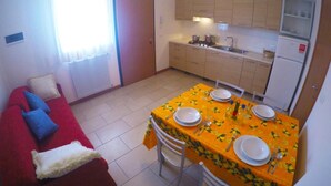 A compact living space with a dining area, kitchenette, and a red sofa with blue cushions. - Residence Le Piramidi (Caorle)