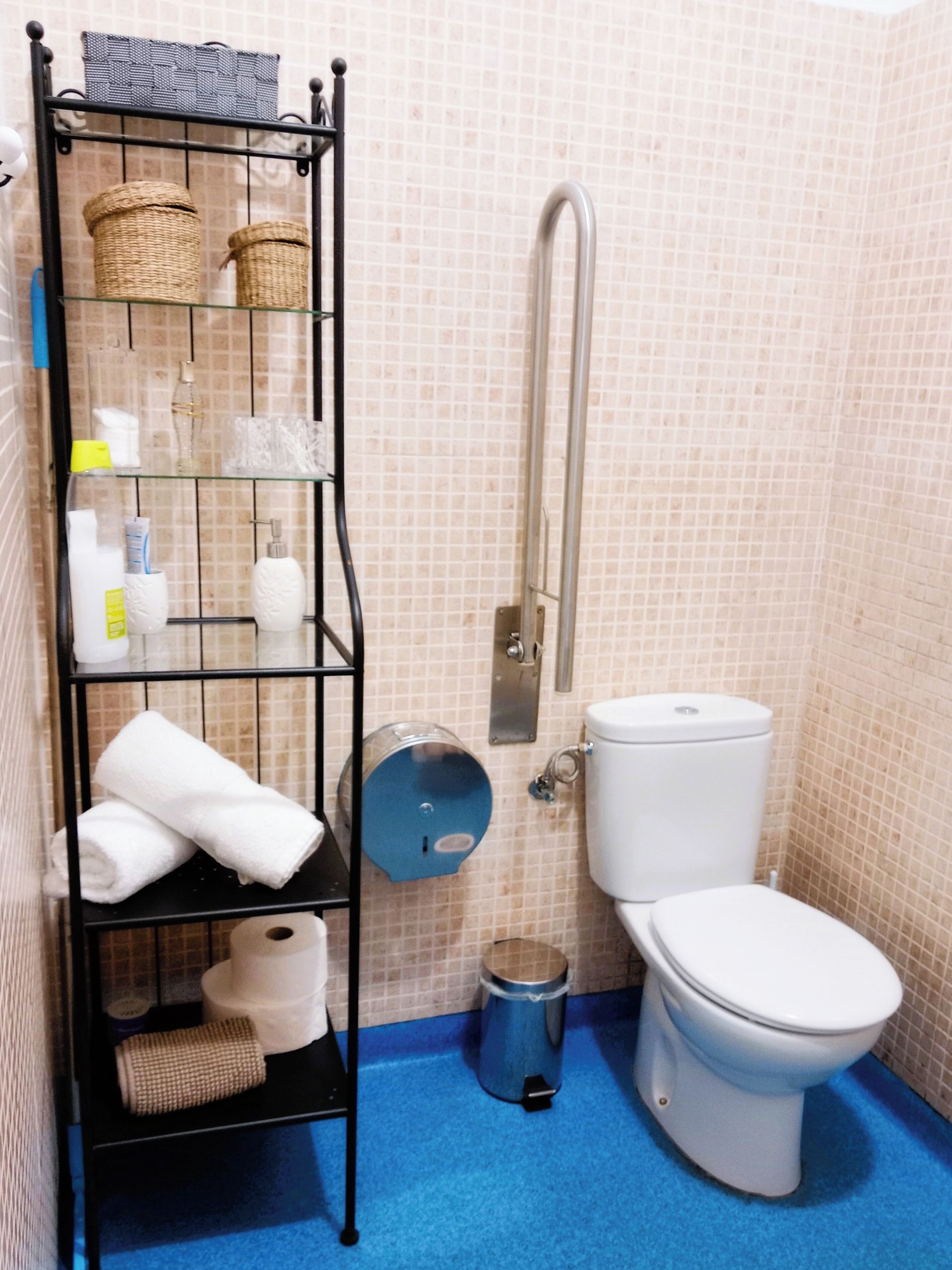 Un baño con un inodoro blanco, piso azul, repisas metálicas con cestas y artículos de aseo, y una zona de ducha.