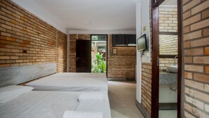 A hotel room with a brick wall, a bed with white linens, a television, and a bathroom with a sink and mirror. - Economy Flat (Natal)