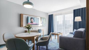 Una habitación de hotel con una mesa de comedor redonda, dos sillas, un sofá, un televisor montado en la pared y un ventanal con cortinas.