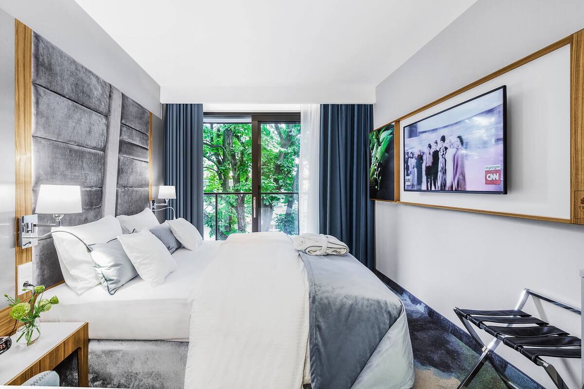 A modern hotel room with a large bed, a TV mounted on the wall, and a window with a view of trees.
