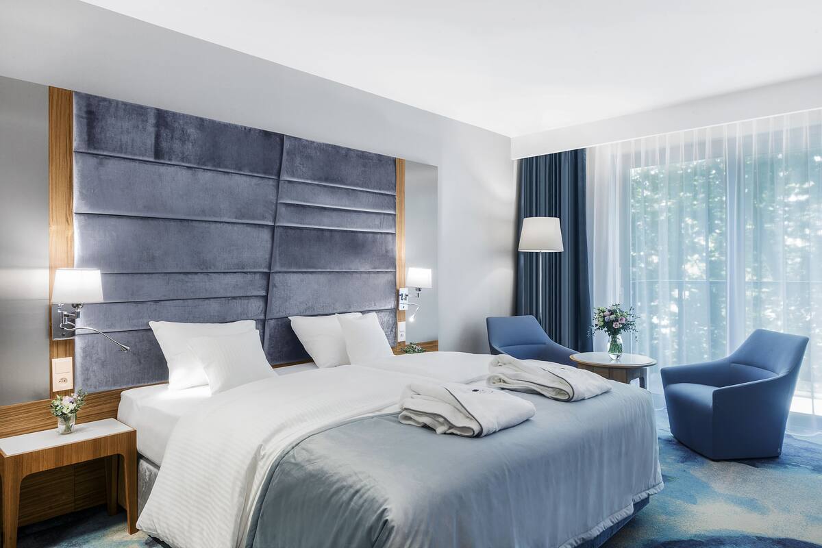 A hotel room with a large bed, two bedside tables, a chair, and a view of trees through the window.