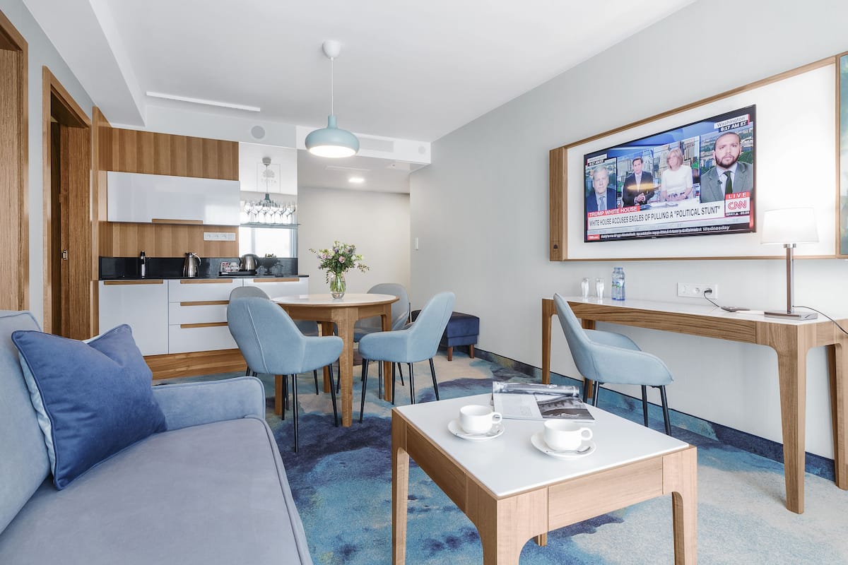 A modern hotel room with a sofa, a dining table with chairs, a small kitchen area, and a TV displaying a news program.
