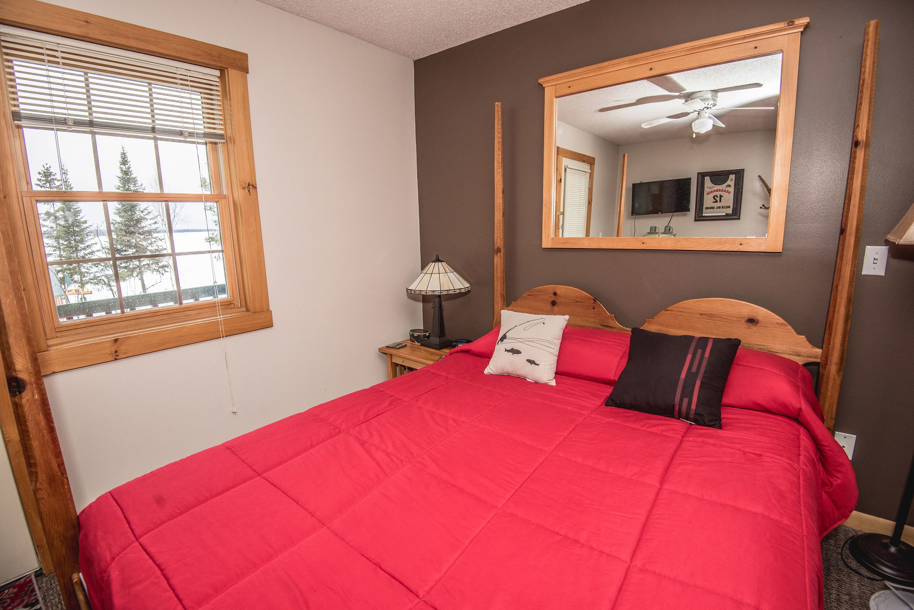 A bedroom with a large bed, a wooden headboard, a nightstand with a lamp, a mirror, and a window with blinds.