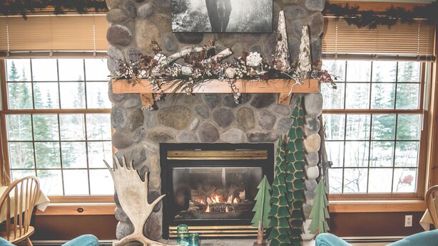 A stone fireplace with a mantel, flanked by two blue armchairs, a wooden floor, and a window with blinds. - Skyport Lodge (Grand Marais)