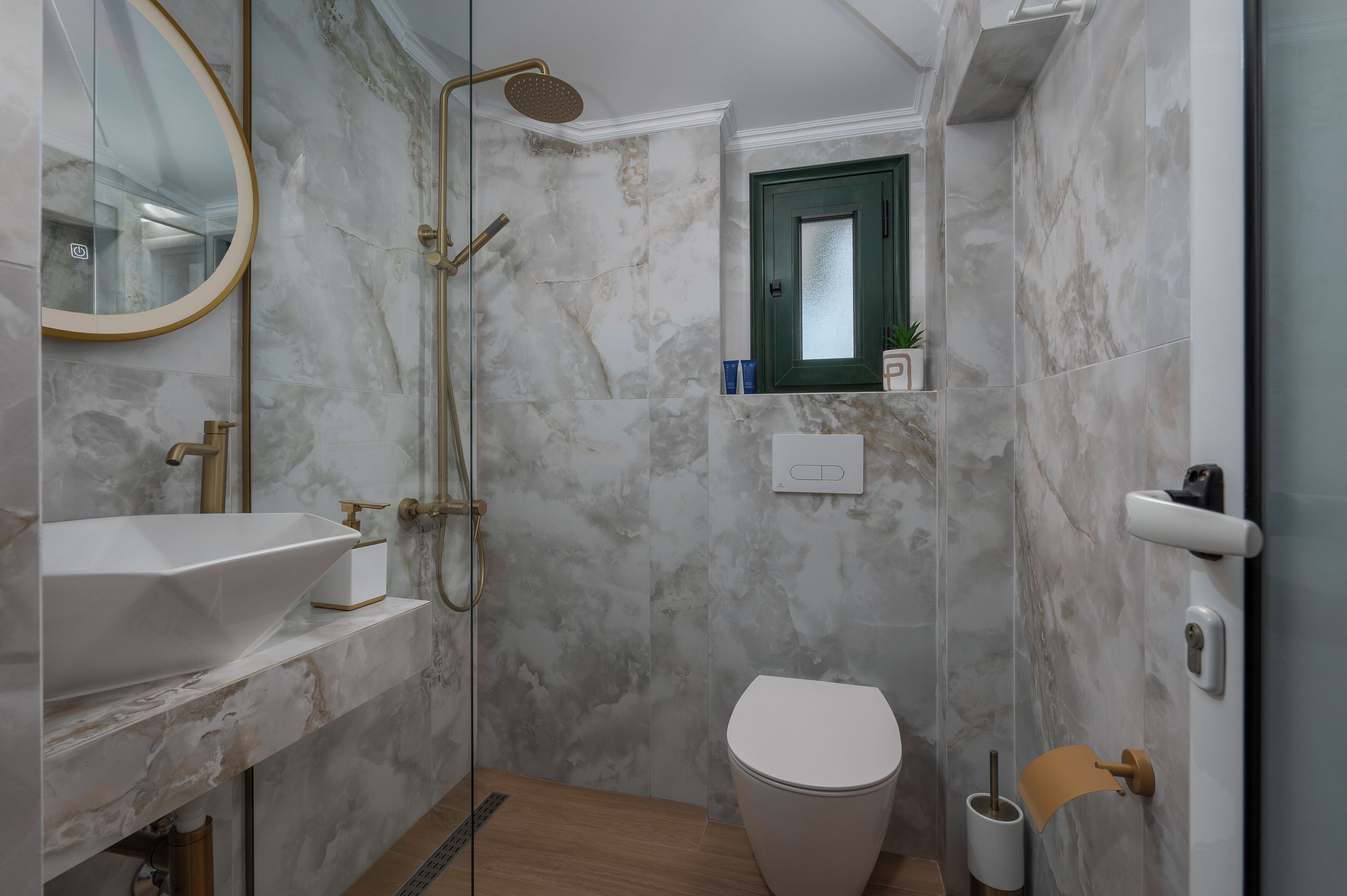 A bathroom with a white sink, a shower with a gold showerhead, a toilet, and a green-framed window.