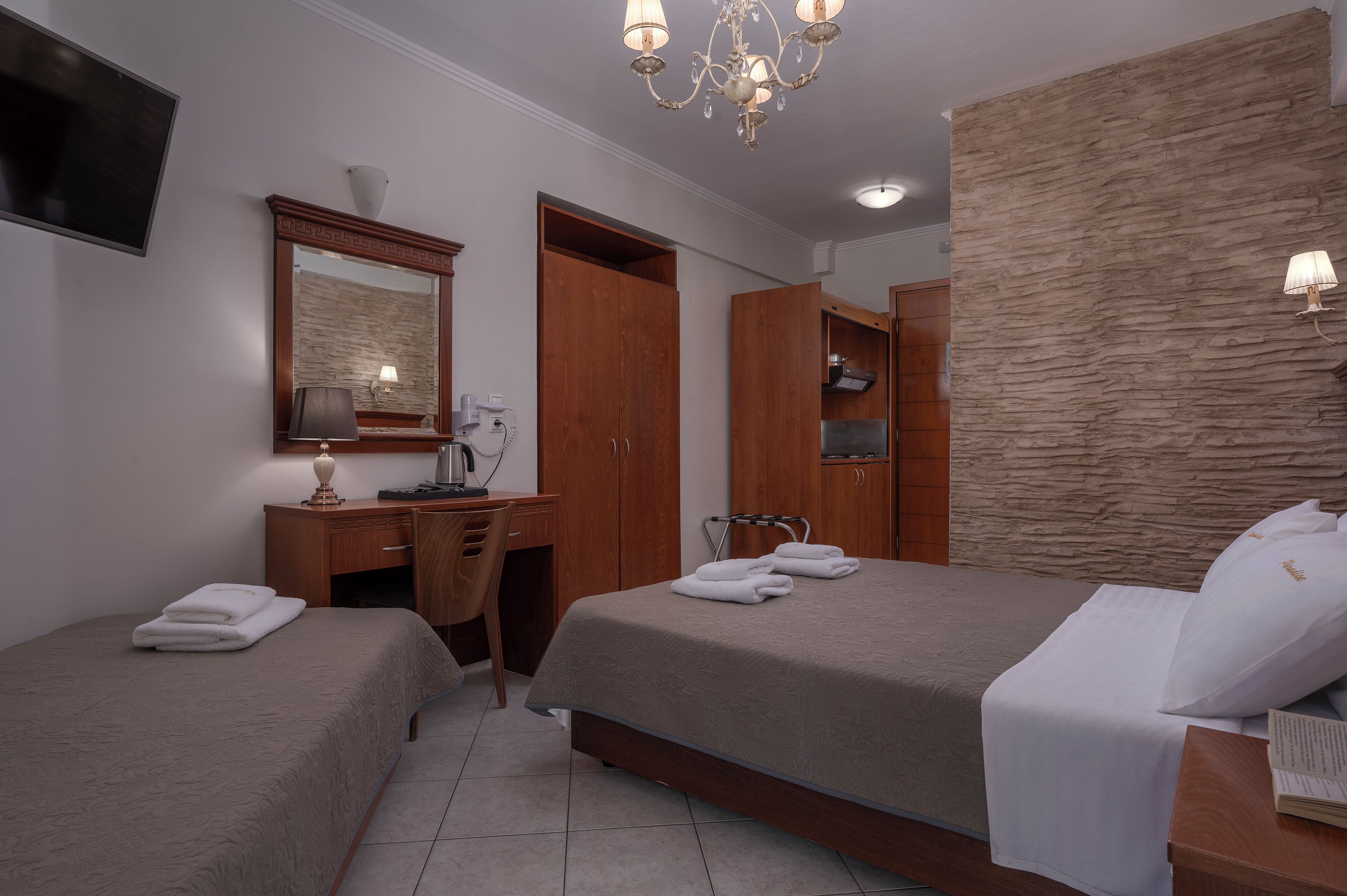 A hotel room with two beds, a desk, a chair, a mirror, a chandelier, and a stone-textured wall.