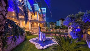 A multi-story hotel with a well-lit outdoor area featuring a water fountain, palm trees, and a stone wall. - Paradise (Volvi)