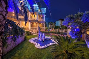 Hotel de varios pisos con un área exterior bien iluminada que cuenta con una fuente de agua, palmeras y un muro de piedra.