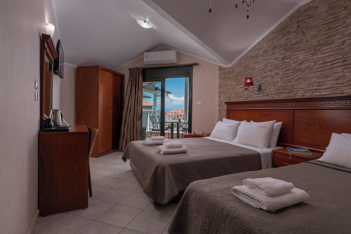 Habitación de hotel con dos camas, un escritorio de madera, un balcón con vistas y una pared de piedra.