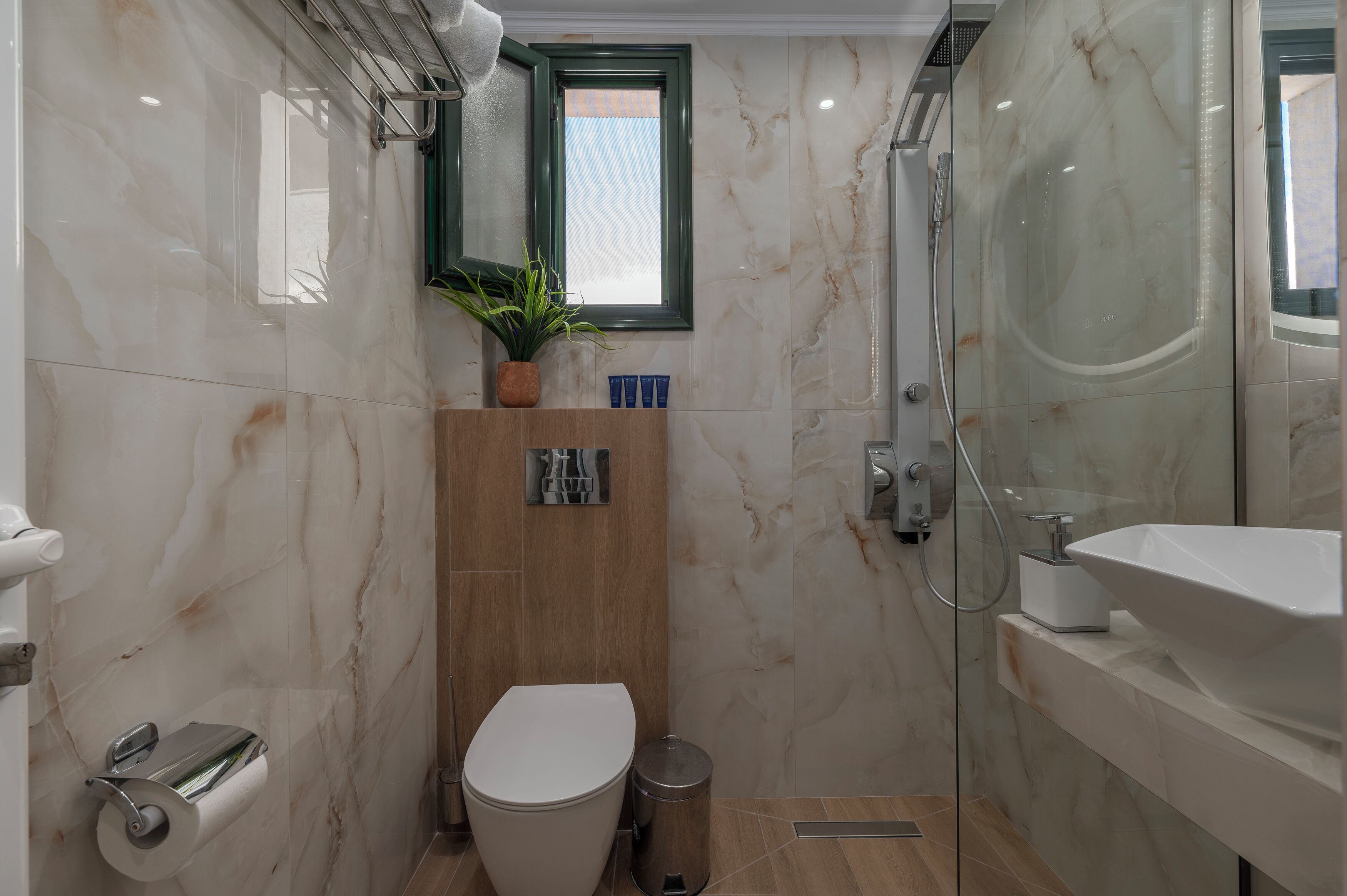 A modern bathroom with a marble wall, a wooden vanity, a white sink, and a glass-enclosed shower.