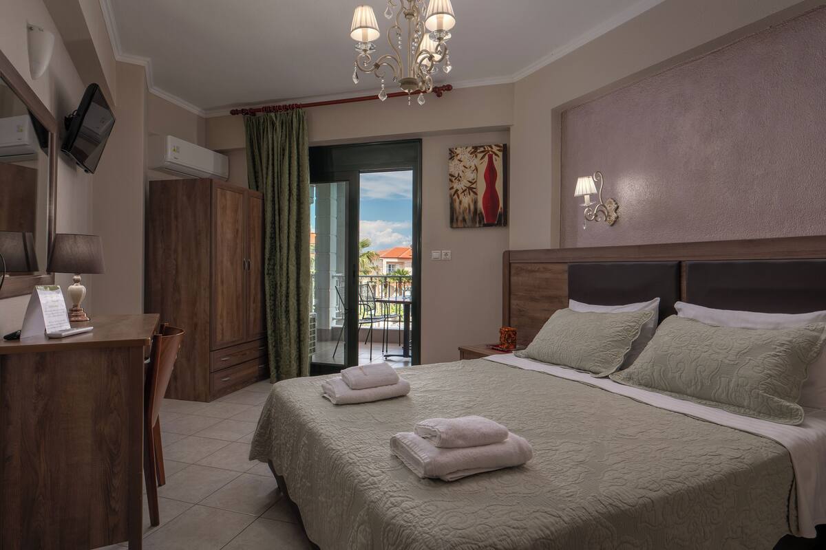 Habitación de hotel con cama, escritorio, silla, televisor y balcón con vistas.