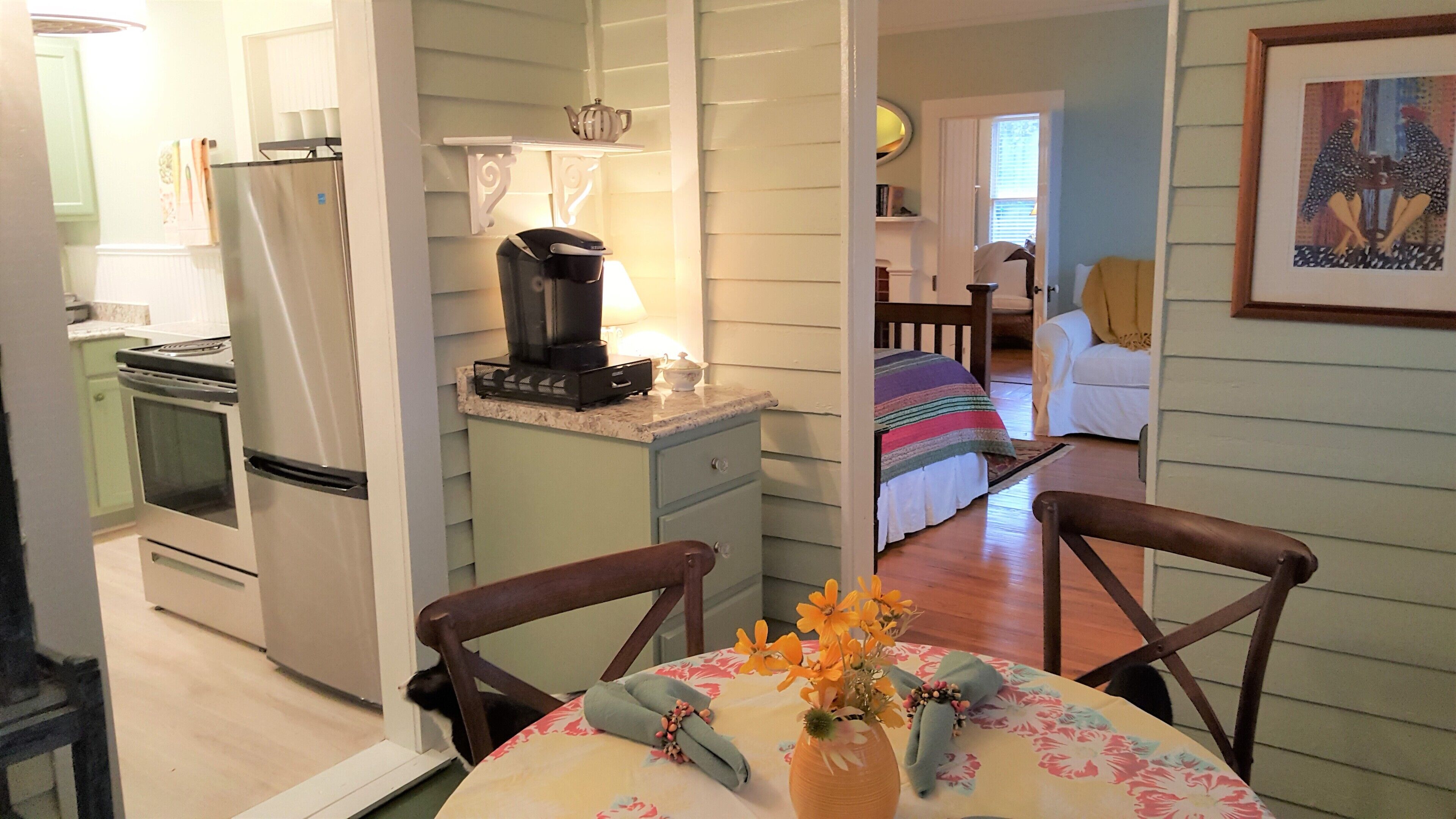 A compact kitchen with a refrigerator, stove, and microwave. There is a dining table with chairs and a vase with flowers. A bed with a striped blanket is visible in the background.