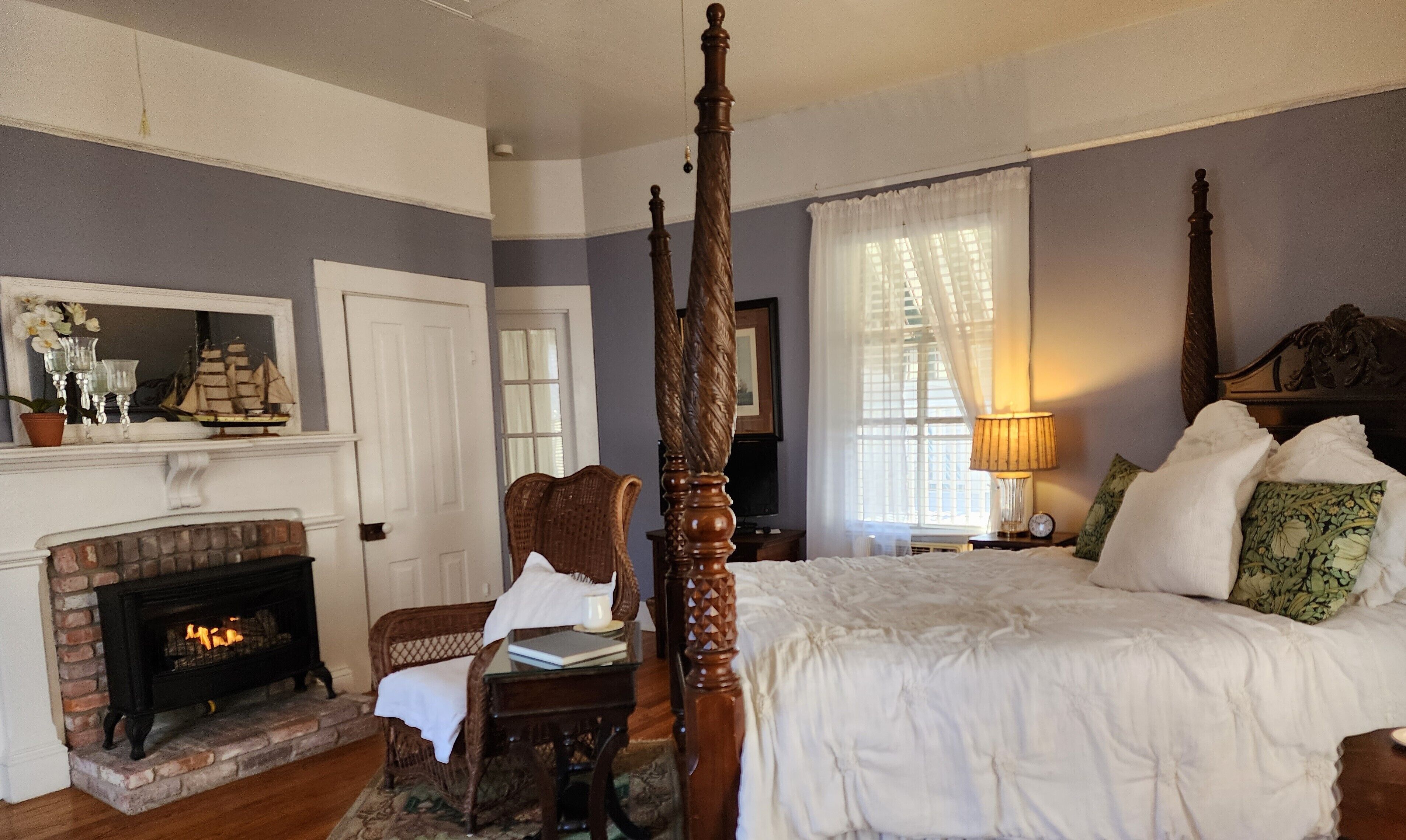 A bedroom with a four-poster bed, a fireplace, a wicker chair, and a nightstand with a lamp.