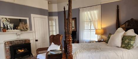 A bedroom with a four-poster bed, a fireplace, a wicker chair, and a nightstand with a lamp.