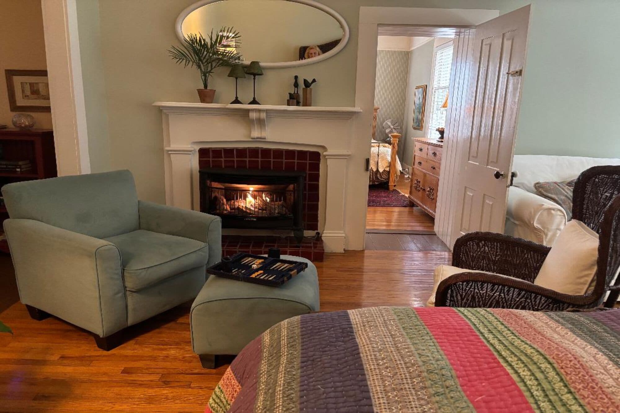 A cozy living room with a fireplace, a comfortable armchair, a sofa, and a bed with a colorful quilt.