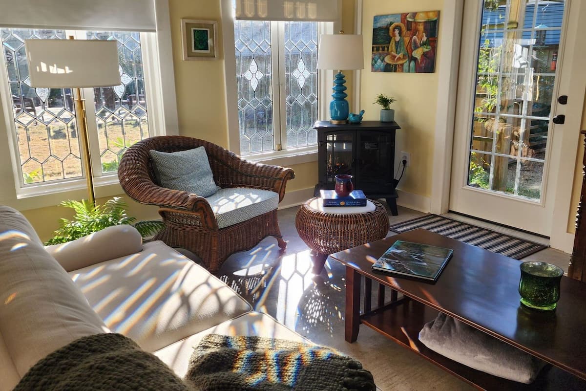 A cozy living room with a wicker armchair, a wooden coffee table, and a sofa. There is a fireplace with a blue vase and a book on the mantel.