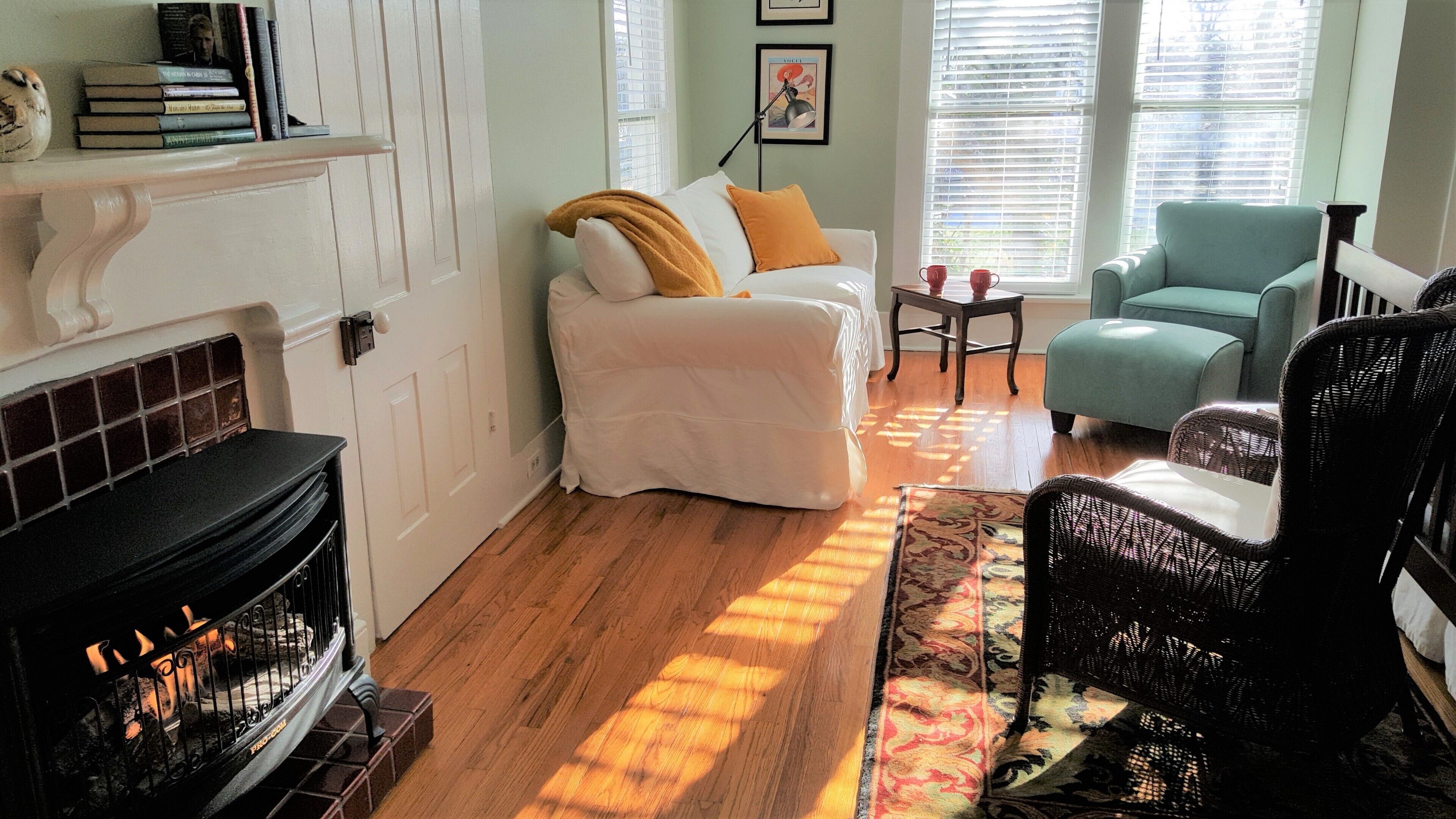 A cozy living room with a fireplace, a white sofa with a mustard throw, a teal armchair, and a wicker chair.