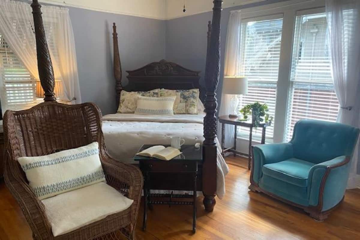 A bedroom with a four-poster bed, a wicker chair, a blue armchair, and a bedside table with a book.